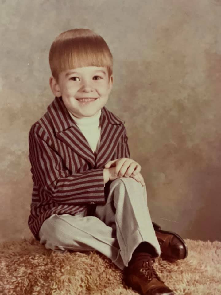Lance about age 7 posing on shag carpet with an indistinct photo backdrop with a bowl hair cut and wearing a white turtleneck with a red and gray striped jacket, tan pants and shiny brown dress shoes.