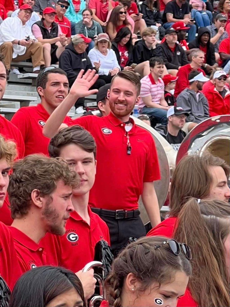 Barron Wallace in his red University of Georgia polo shirt waves at the camera in the middle of the Redcoat band in the stands at Sanford Stadium in Athens during the spring game in April 2022.